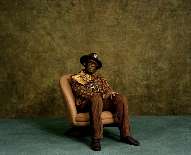 Mature African American man poses in an armchair wearing a hat, patterned jacket and scarf in front of a textured backdrop.