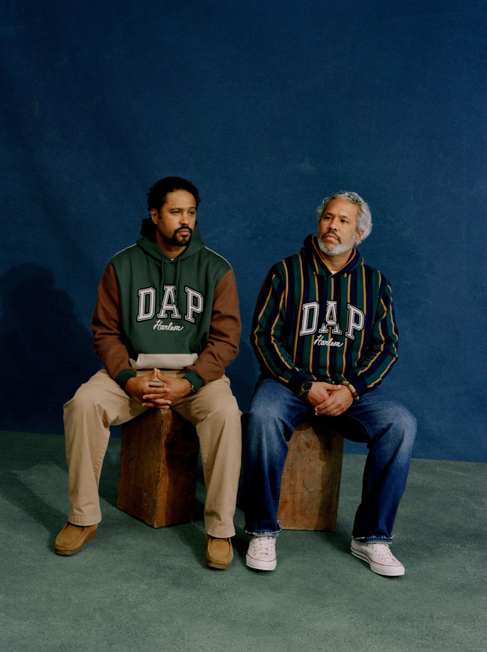 Two adult men in hooded sweatshirts sit side-by-side, wearing blue jeans and canvas shoes against a textured blue backdrop.