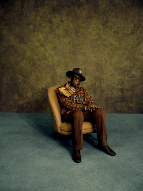 Man in patterned jacket and fedora sits in beige chair against mottled backdrop. Brown patterned pants and shoes. Neutral tones.