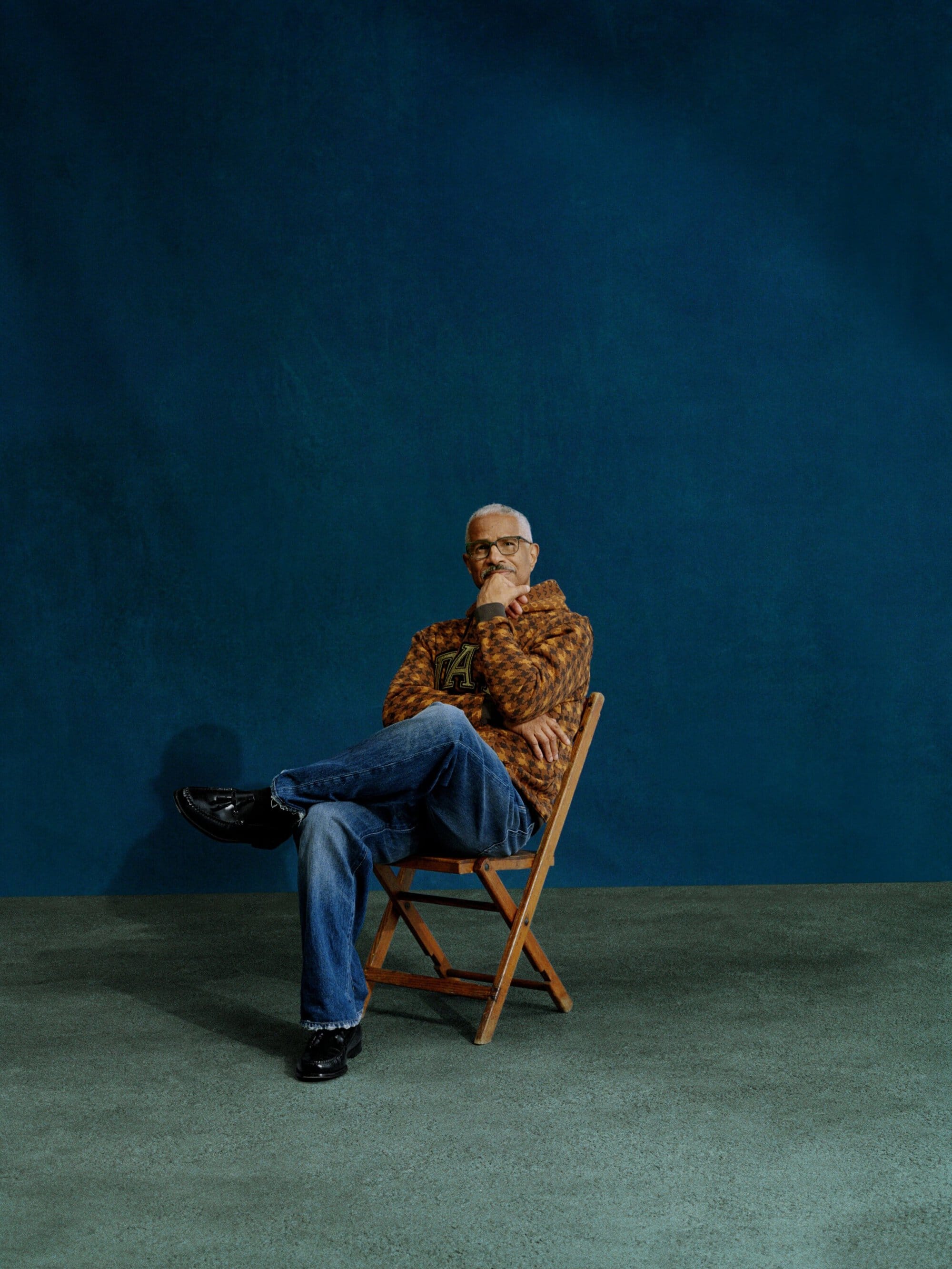 Portrait of a mature man with salt and pepper hair, wearing a patterned hoodie and jeans, sitting on a wooden chair against a blue backdrop.