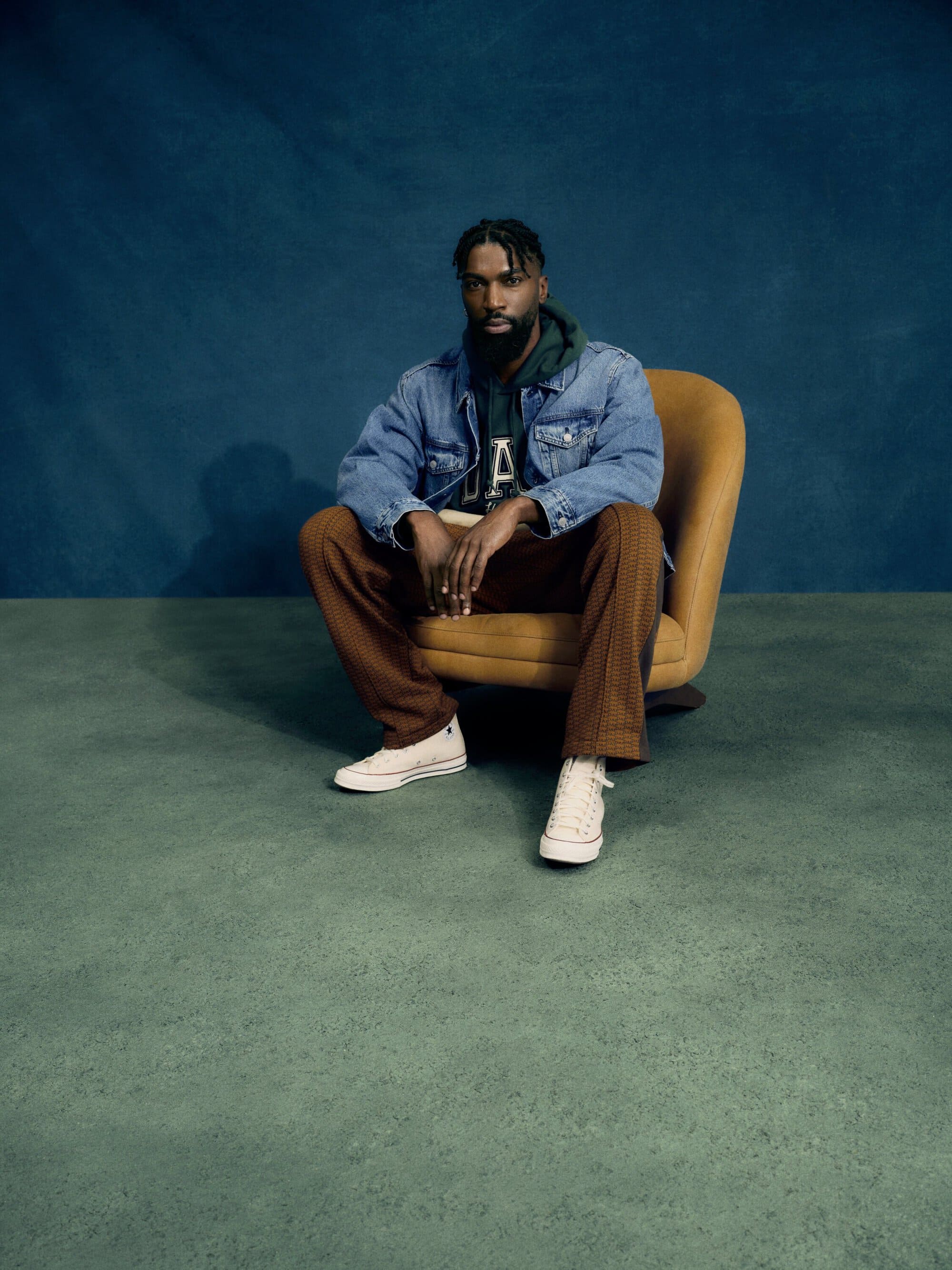 Portrait of a Black male seated in a yellow chair, wearing a denim jacket, plaid pants and sneakers against a blue and green background.