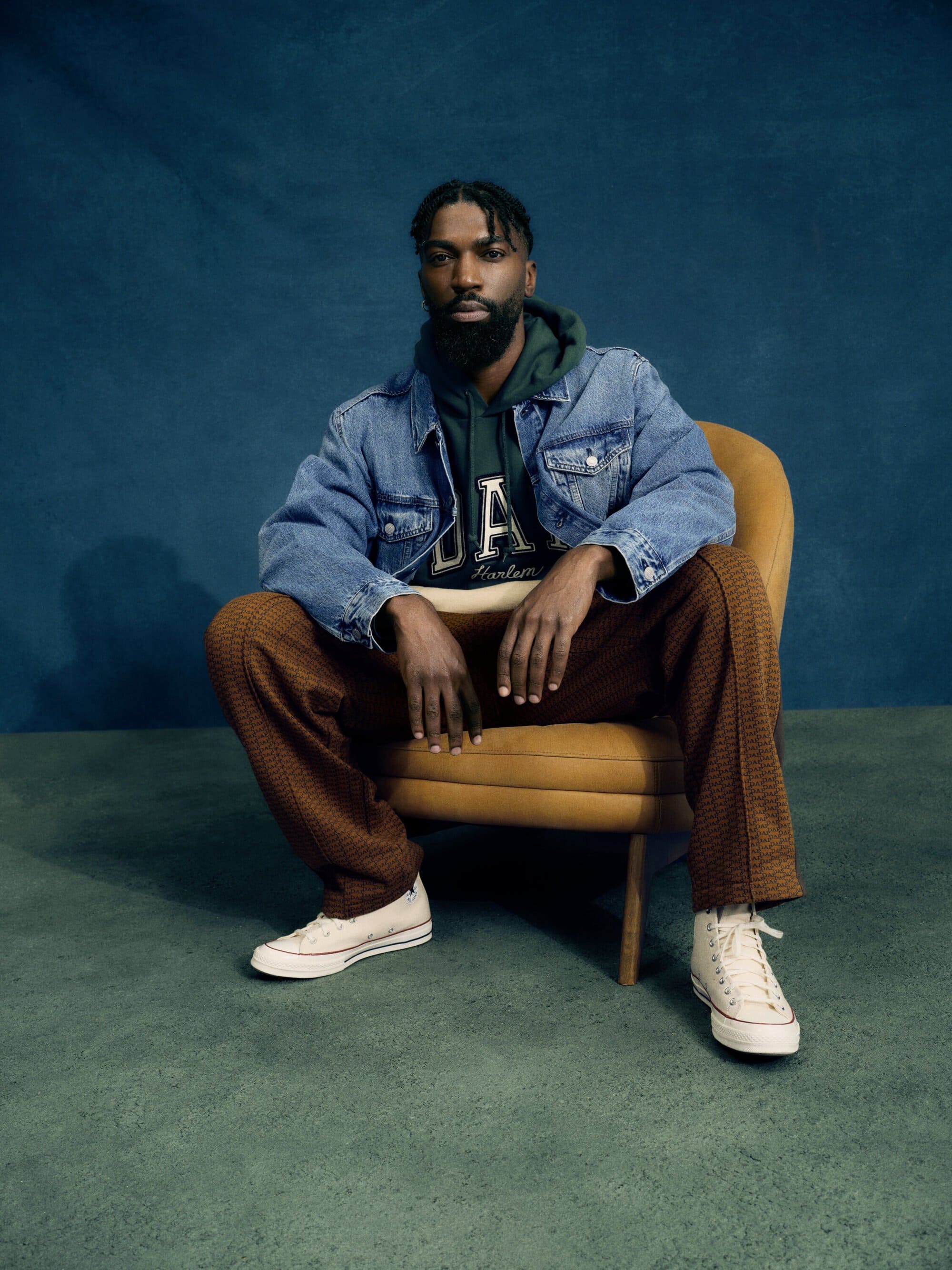 A bearded Black man in a denim jacket and sweat suit poses sitting on a yellow chair against a blue background in a studio shot.