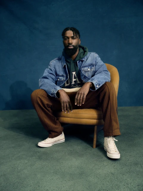 A bearded Black man in a denim jacket and sweat suit poses sitting on a yellow chair against a blue background in a studio shot.