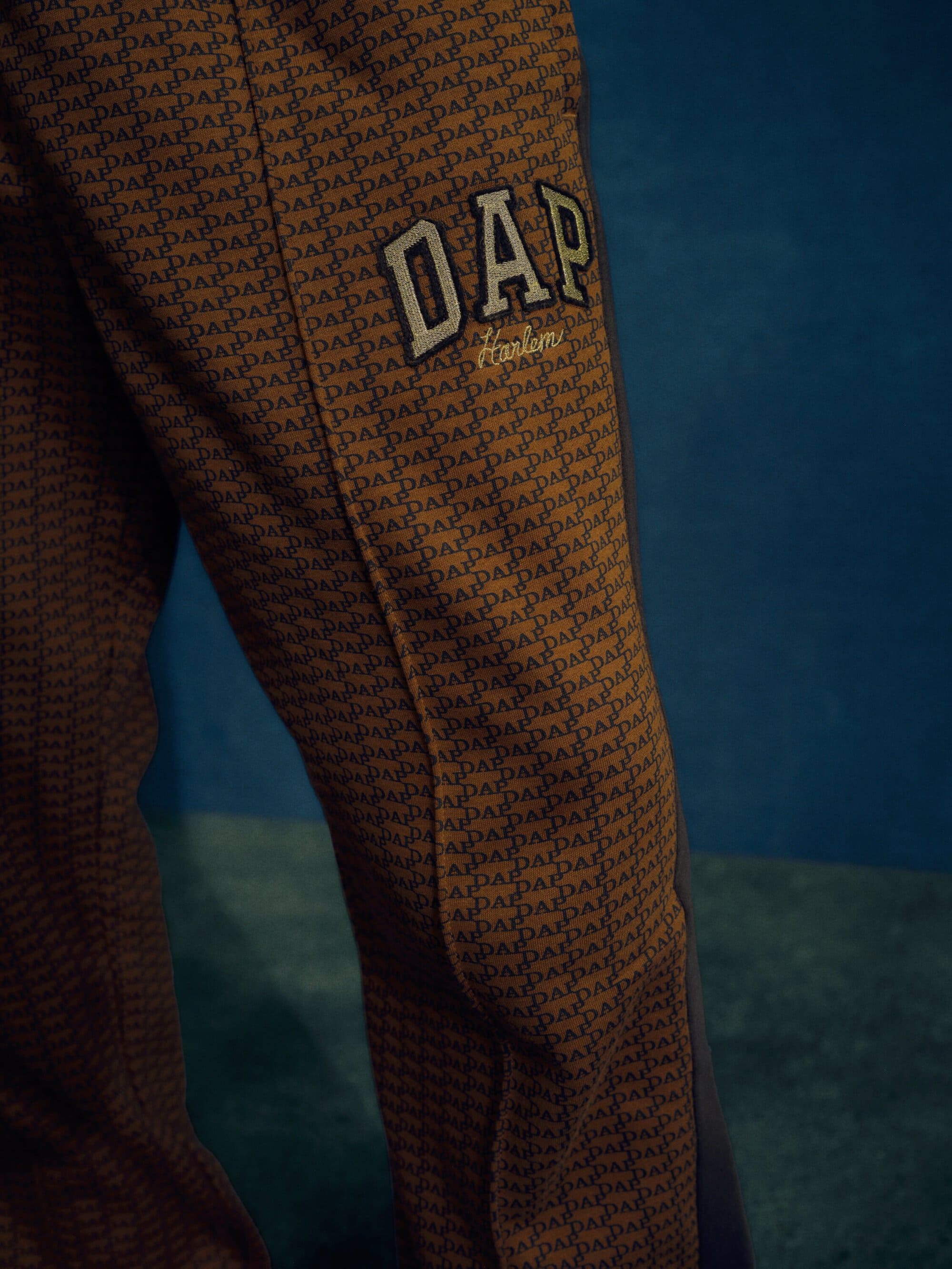 Close-up of patterned brown pants featuring the stitched 'DAP' logo in white against a dark blue background with side panel detailing.