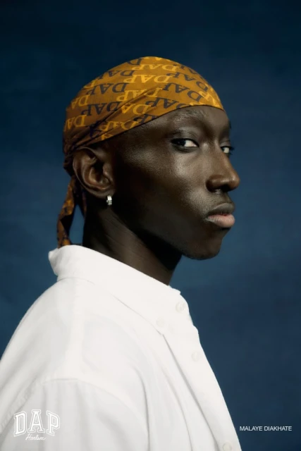 Close-up portrait of a dark-skinned adult male wearing a patterned headscarf and white collared shirt against a dark blue backdrop.