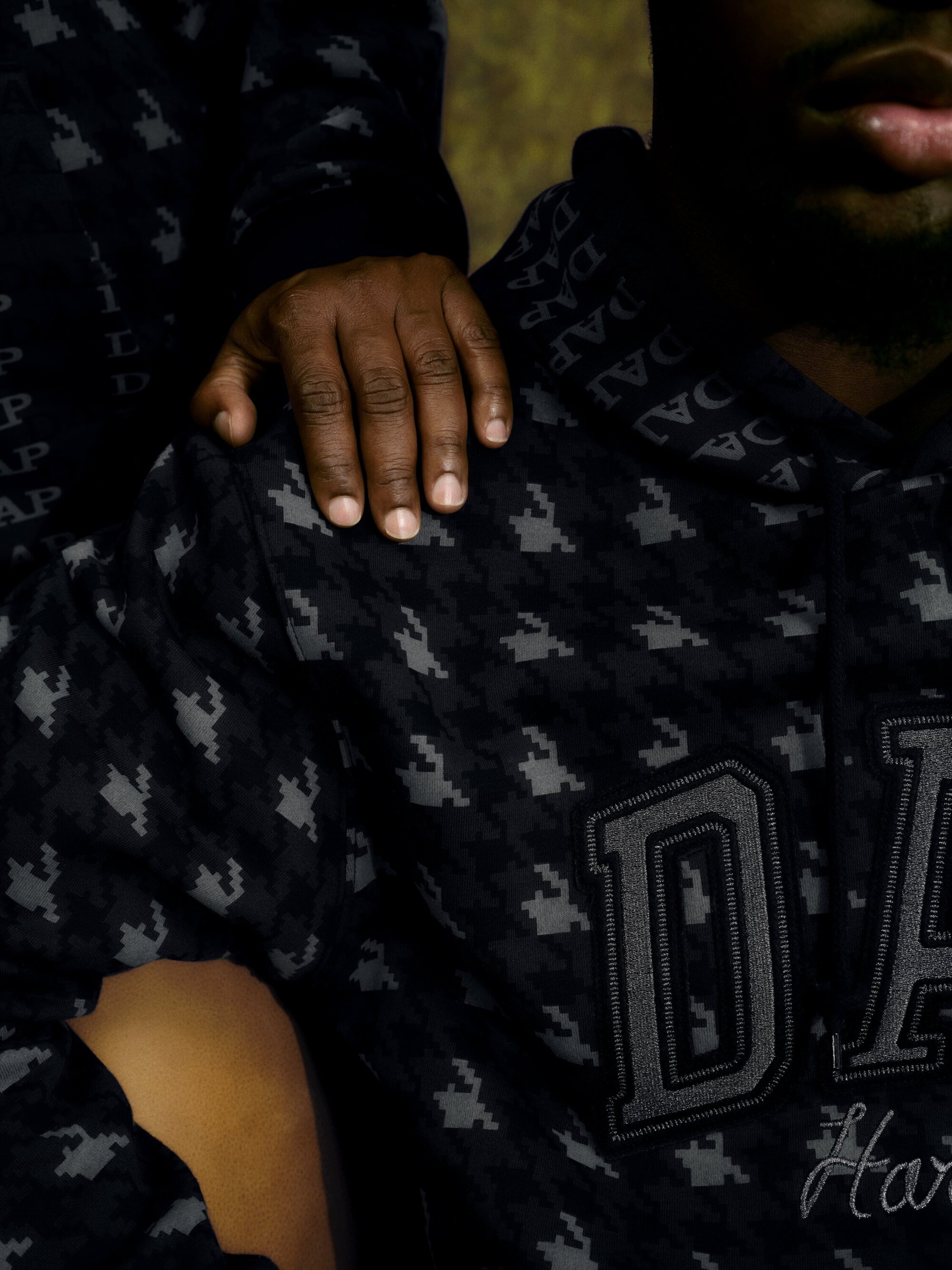 A close-up of two people in patterned hooded sweatshirts, one person's hand is on the shoulder of the other, against a blurred background.