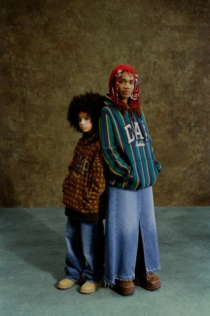 Two young models pose indoors against a mottled backdrop, wearing casual clothing: graphic hoodies, wide-leg jeans, and platform shoes.