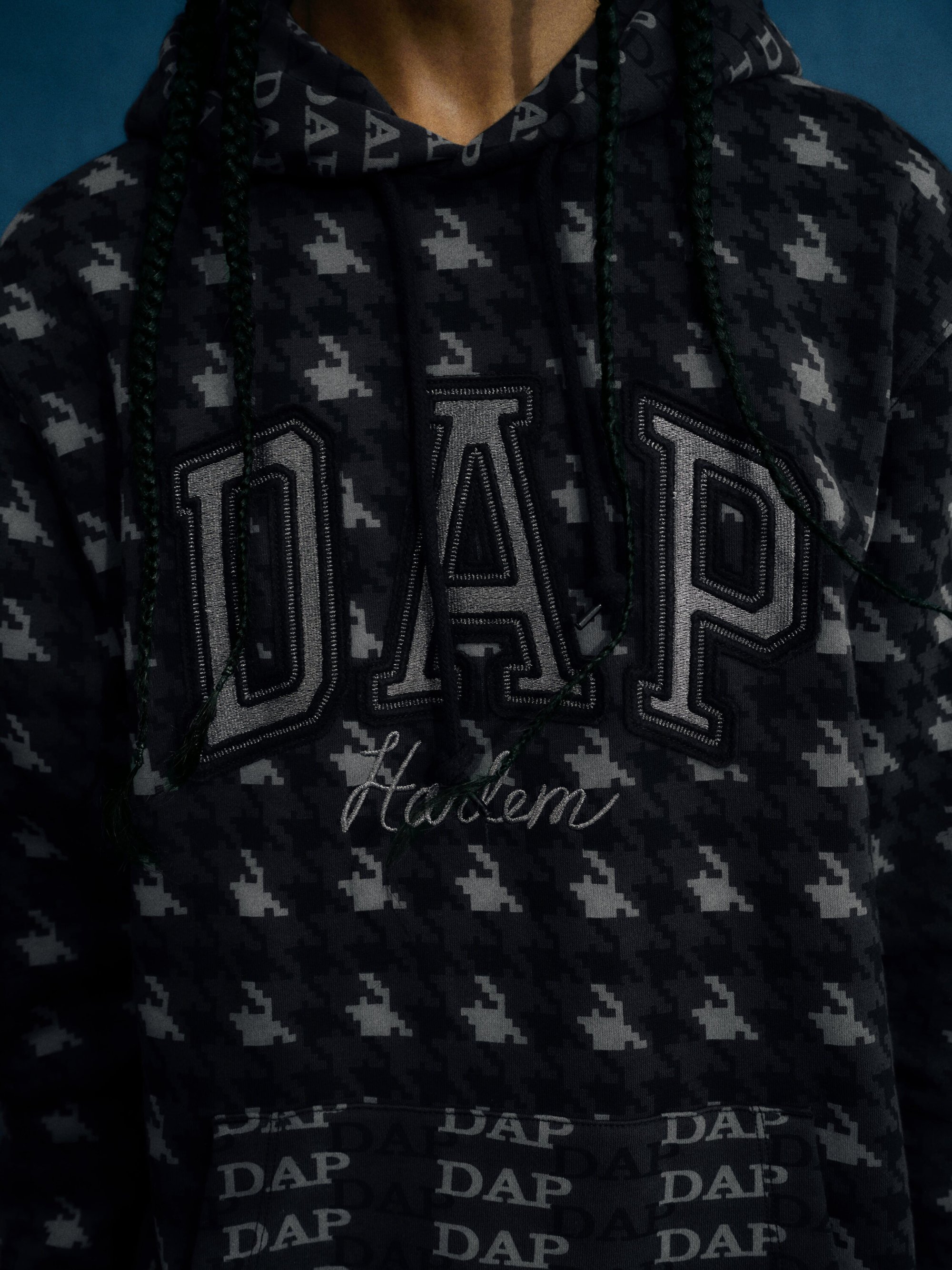 Close-up of a person wearing a black DAP hoodie with a houndstooth pattern and the word 'Harlem' embroidered.