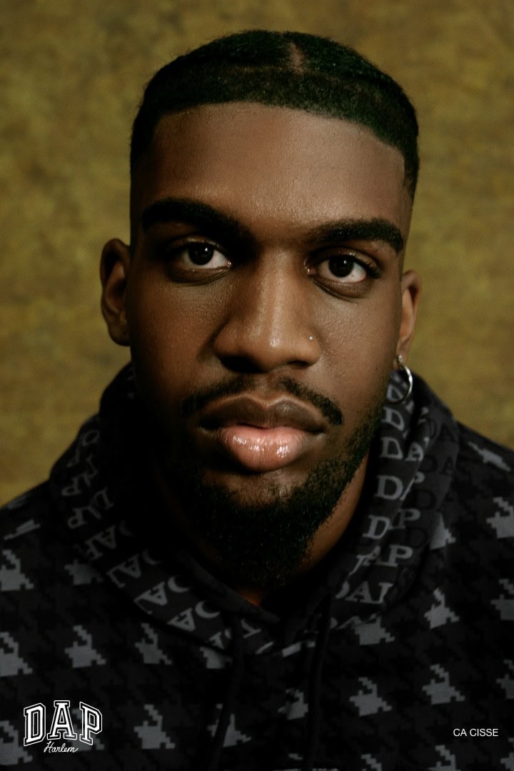 Portrait of a stylish adult male with dark hair and a patterned hoodie against a mottled gold background in an indoor setting.