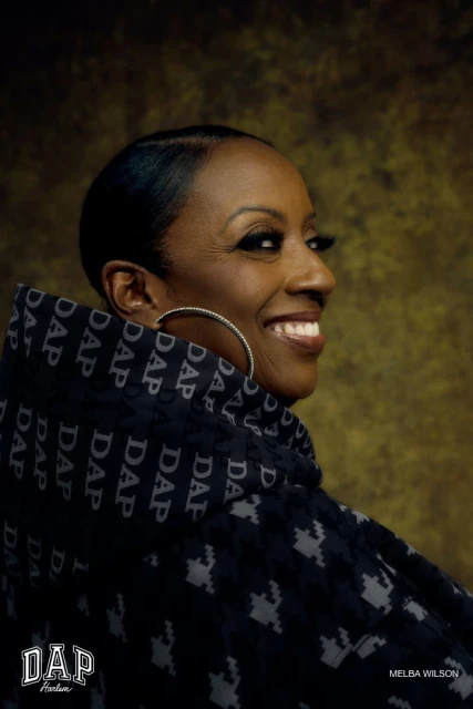 Profile shot of an African-American woman with short black hair, wearing patterned hooded garment, and jewelry, smiling against neutral backdrop.