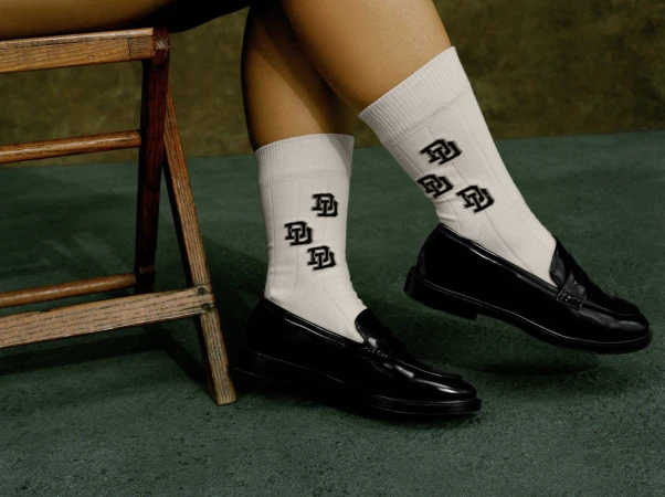 Close-up of legs wearing white branded socks and black loafers next to a wooden stool