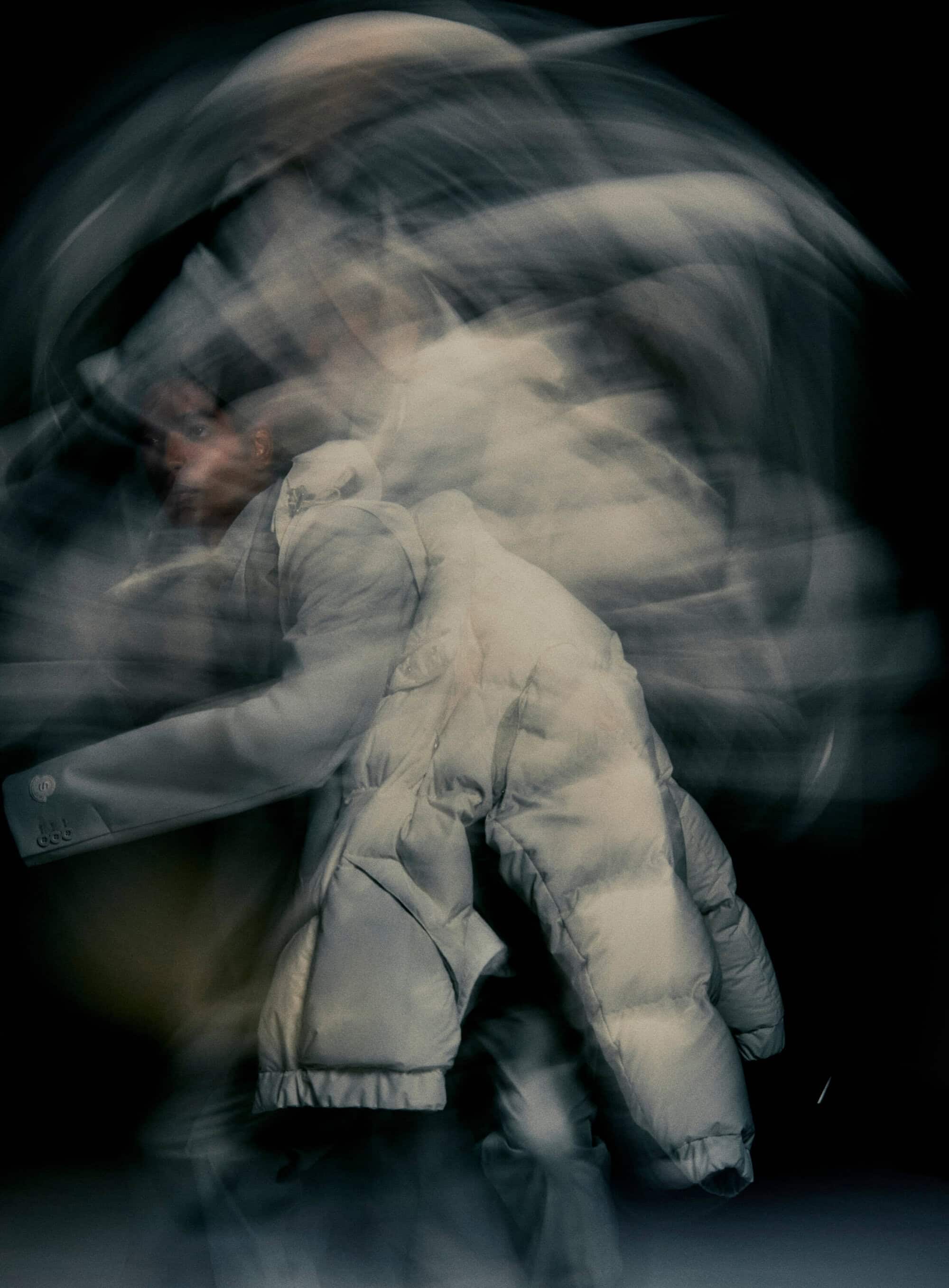 Abstract long exposure of a person in motion wearing a grey puffer jacket against a dark background, creating a dynamic blurred effect.