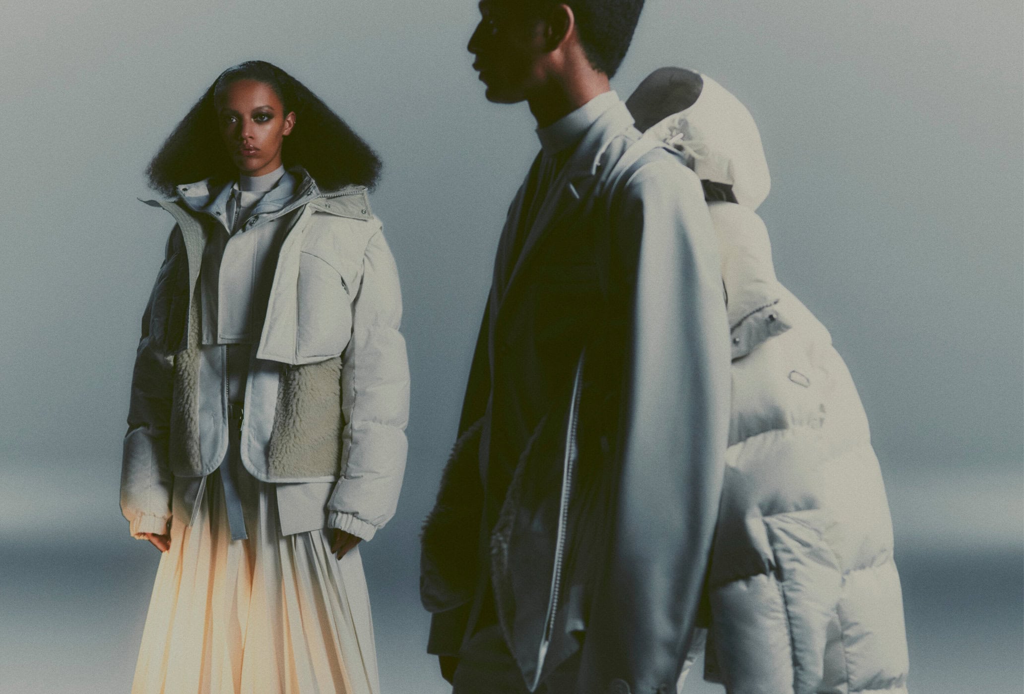 Two people model winter fashion, showcasing puffer jackets and stylish skirts in a studio setting with soft, diffused lighting.