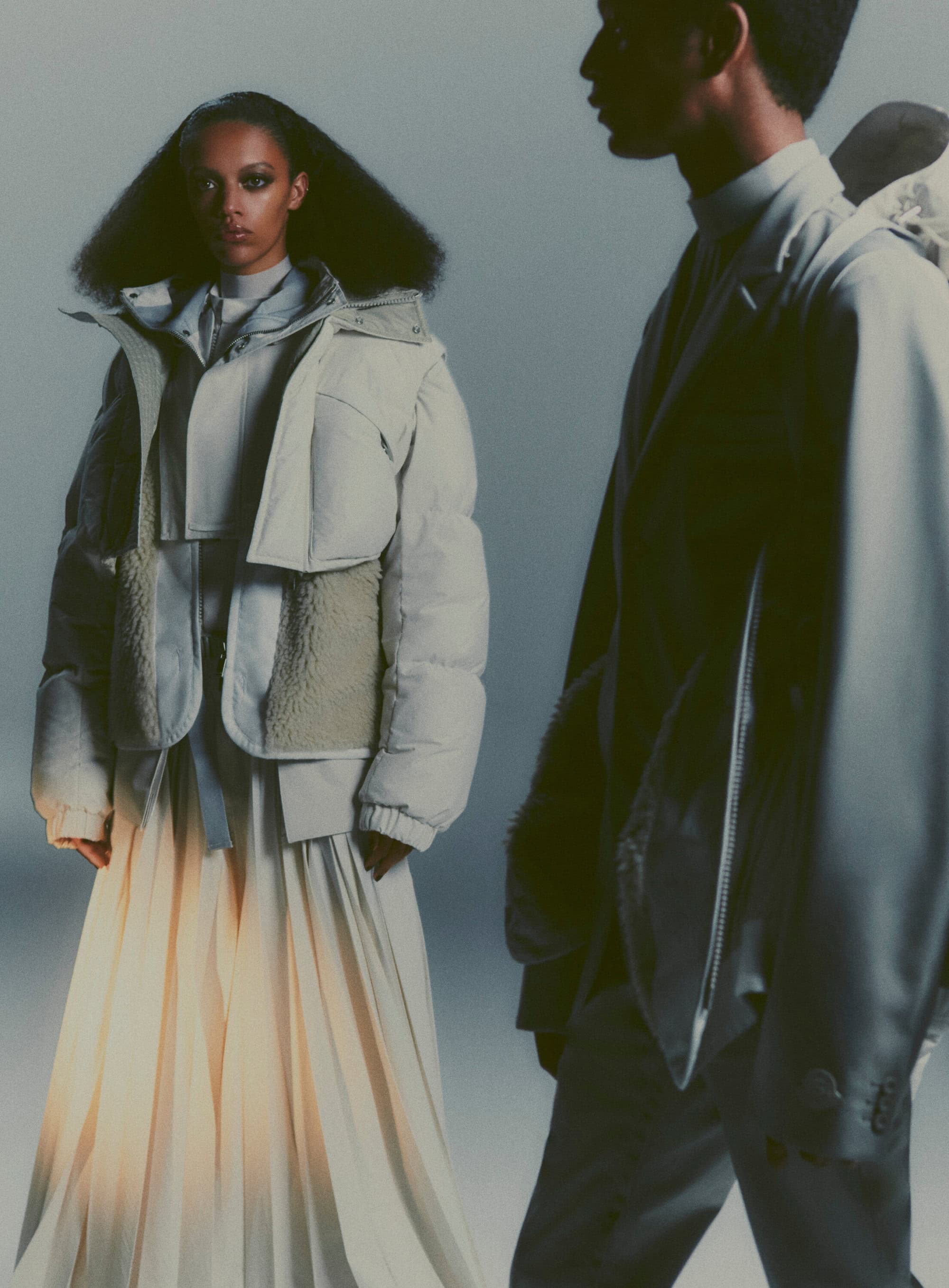 Two fashion models showcase outerwear and formal skirt and jacket against a neutral backdrop in a studio fashion shoot. 