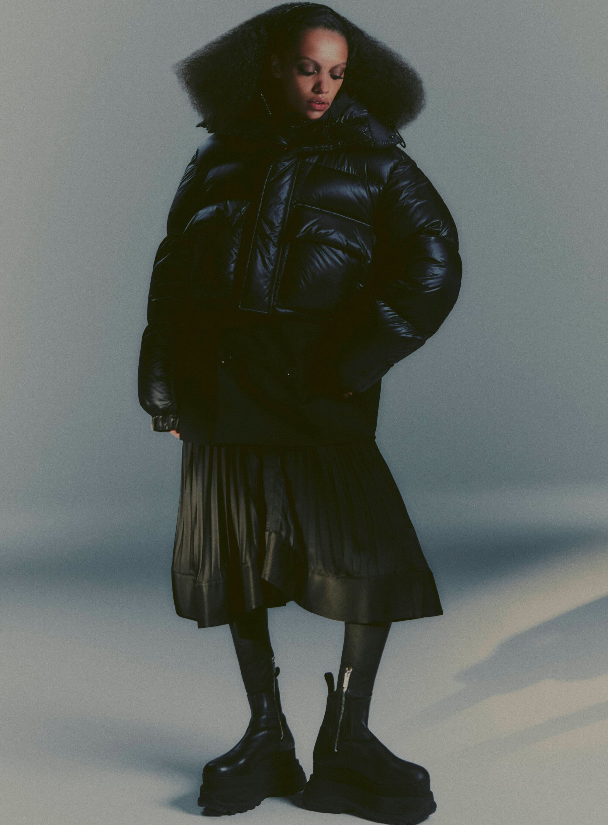 Fashion shot of a woman in a black puffer coat, midi skirt, platform boots against a grey studio backdrop, dramatic and modern style.