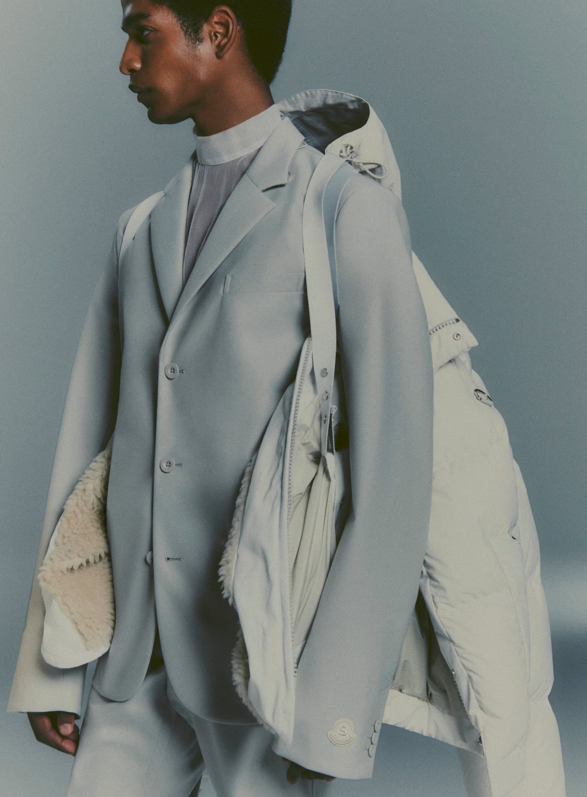 Man in a light grey suit and top stands in front of a neutral background with a white puffer jacket draped on his shoulder.