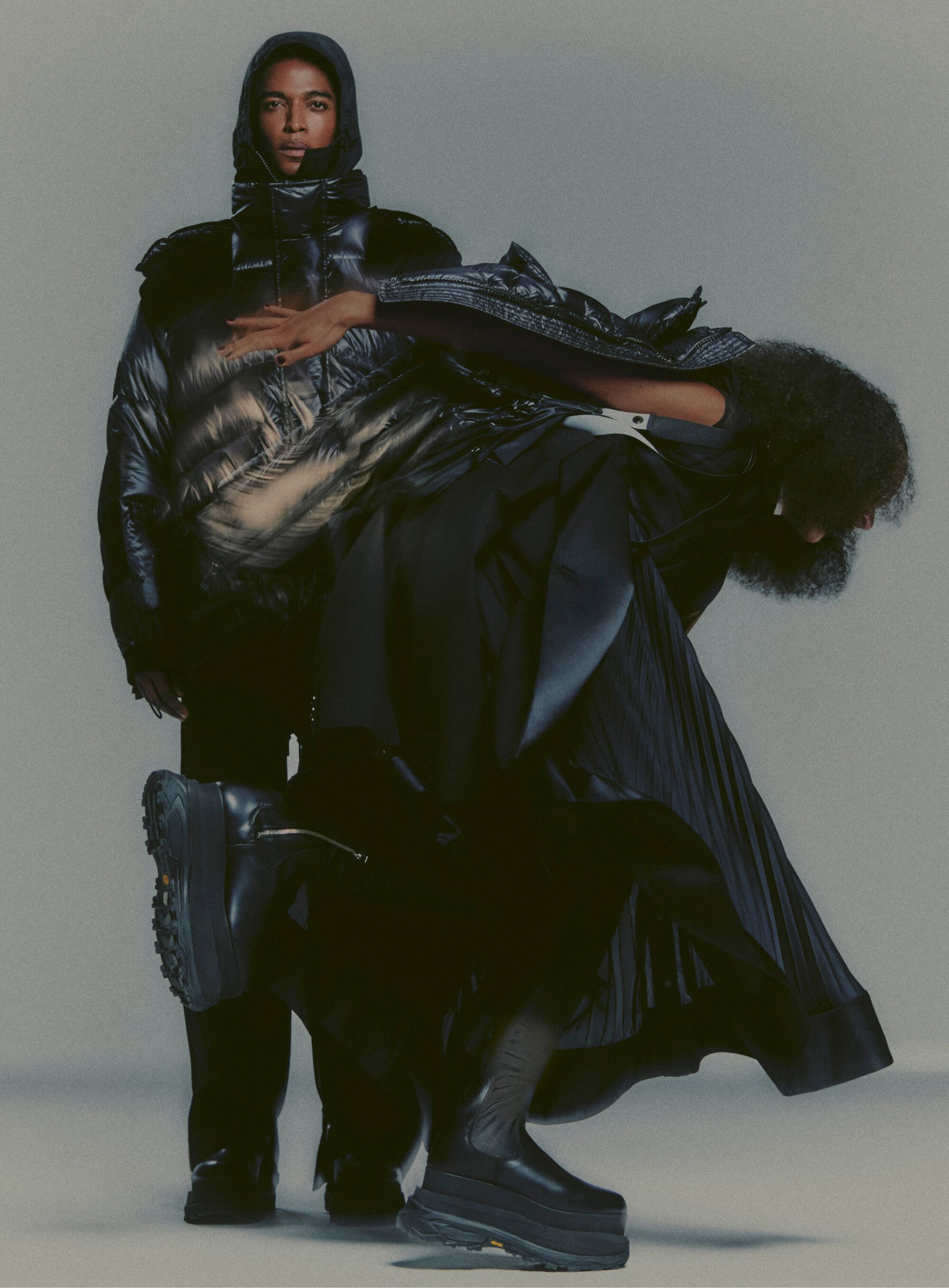 Two people pose wearing dark avant-garde fashion against a neutral background, with one in a hooded puffer jacket and the other with a pleated skirt.