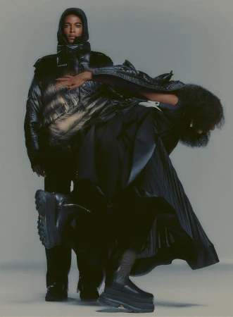 Two people pose wearing dark avant-garde fashion against a neutral background, with one in a hooded puffer jacket and the other with a pleated skirt.