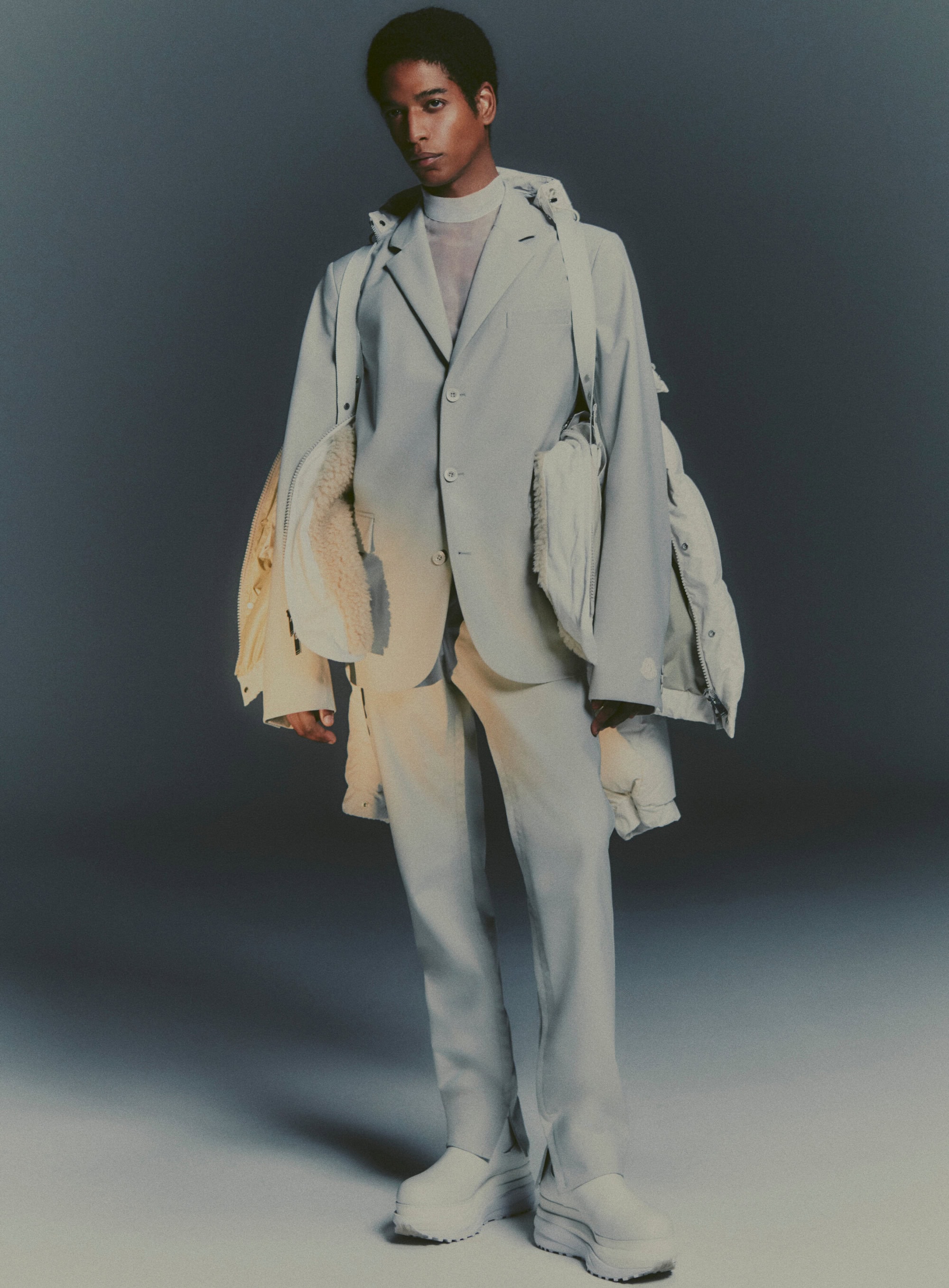 Male model poses in a studio wearing a light-colored suit with a jacket slung over his shoulder, paired with chunky platform shoes.