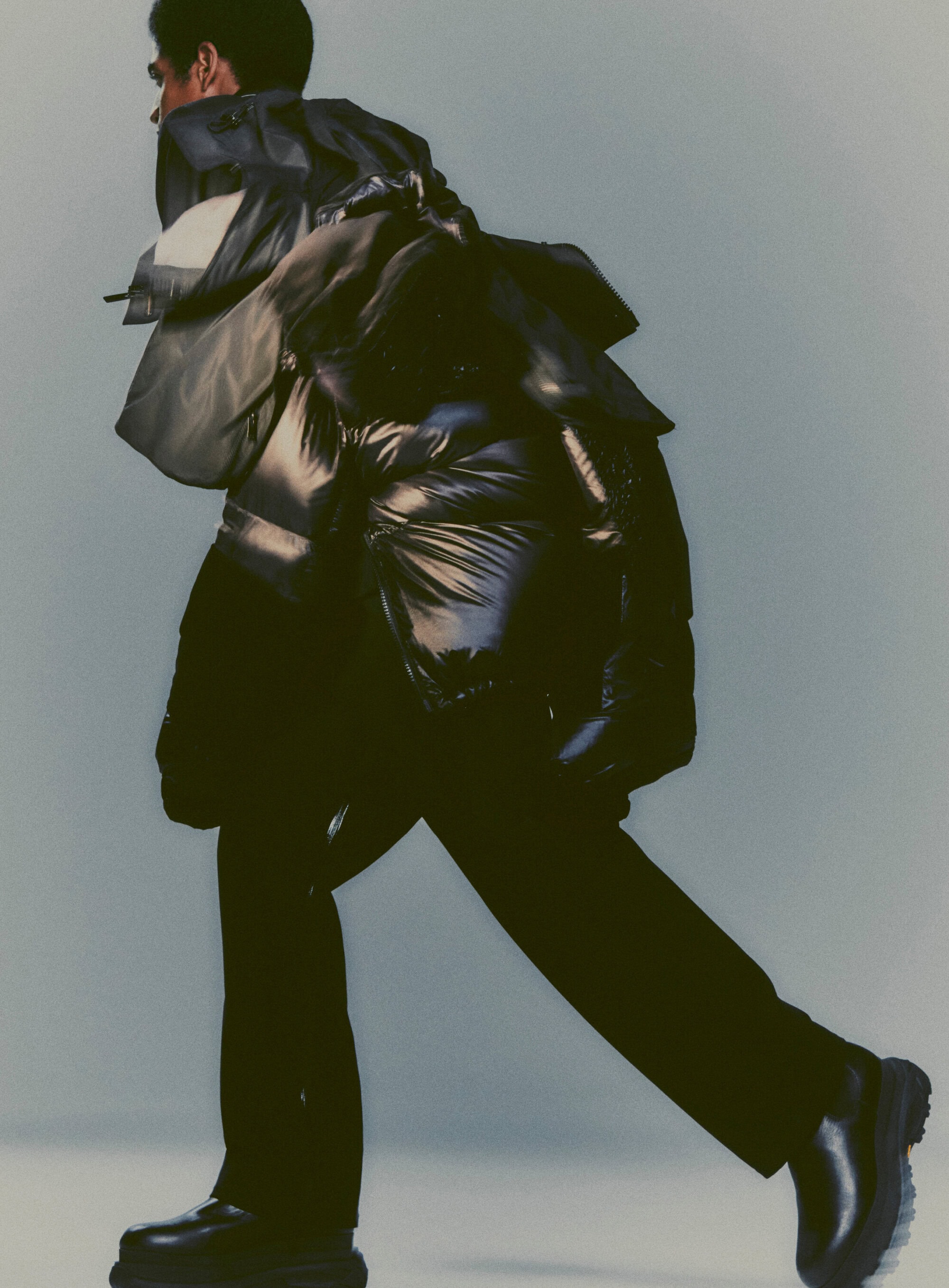 Fashion editorial shot of a person wearing a dark puffer jacket, black trousers, and black boots against a light background in studio lighting.