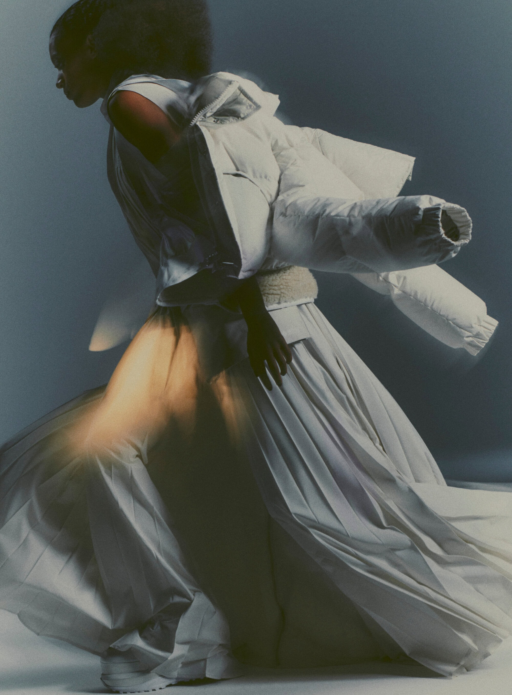Fashion shot of a Black model wearing a white dress and jacket with motion blur, on a neutral background in a photo studio setting.