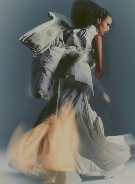 Fashion portrait of a woman with a dynamic pose wearing a futuristic, oversized white jacket and flowing skirt in a studio setting.