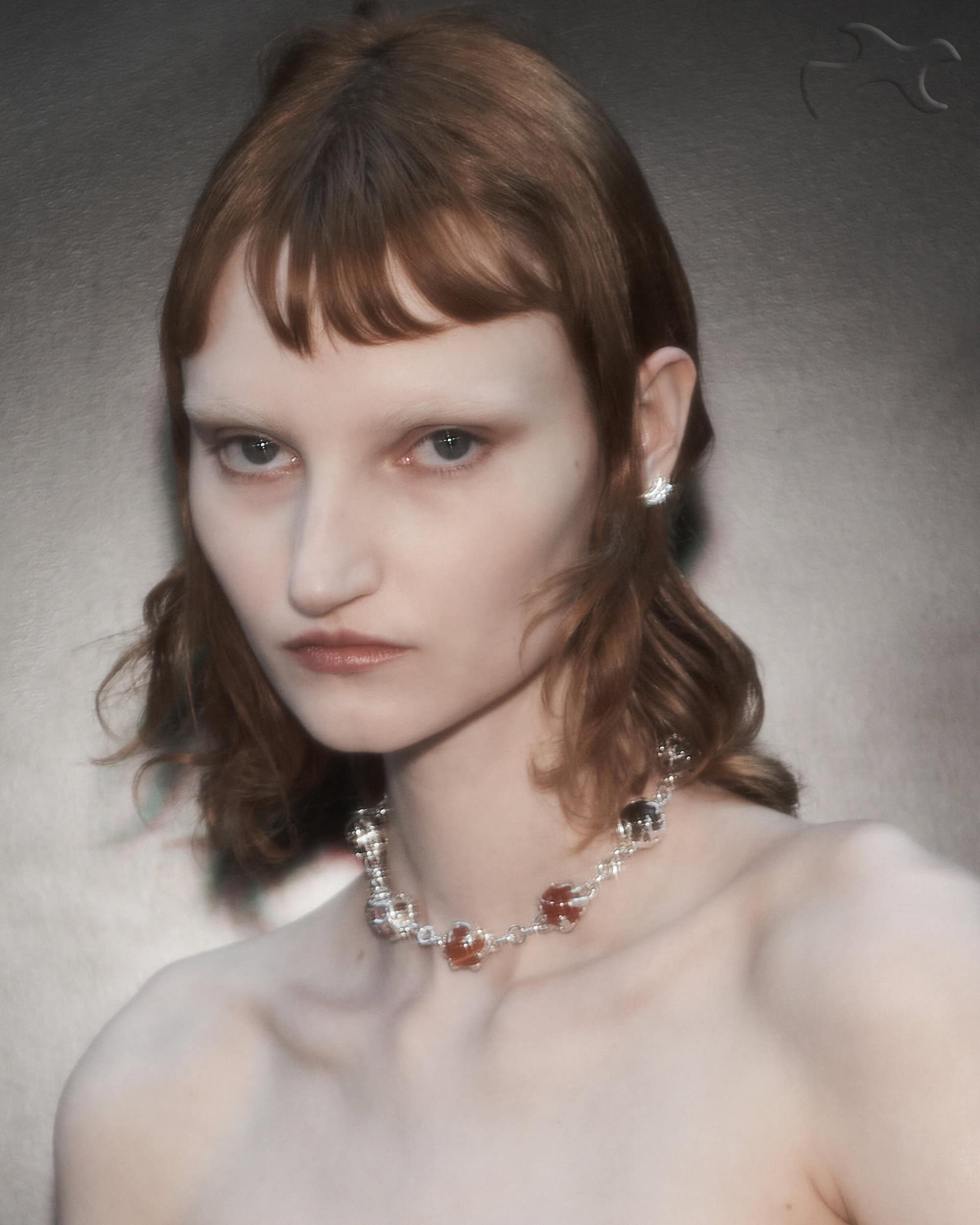 A close-up studio portrait of an auburn-haired person with light skin, wearing a floral necklace and earrings against a dark background.