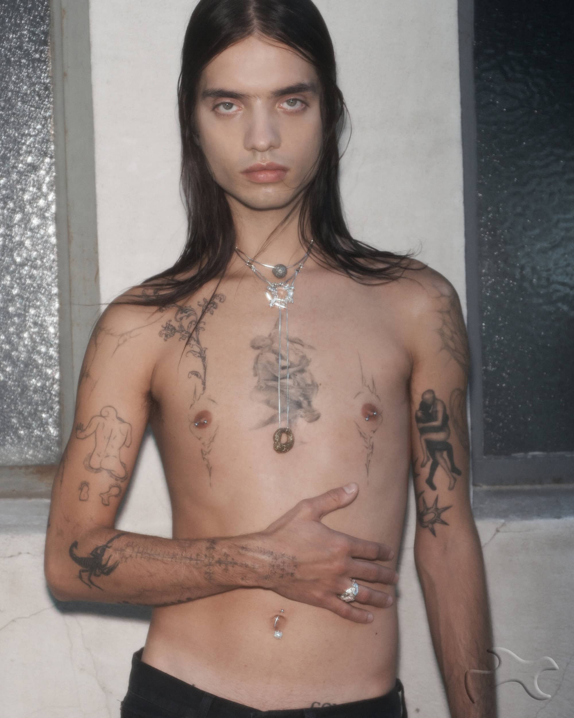 Portrait of a shirtless adult male with long hair, multiple tattoos, and a pendant necklace, posing indoors with a neutral expression.