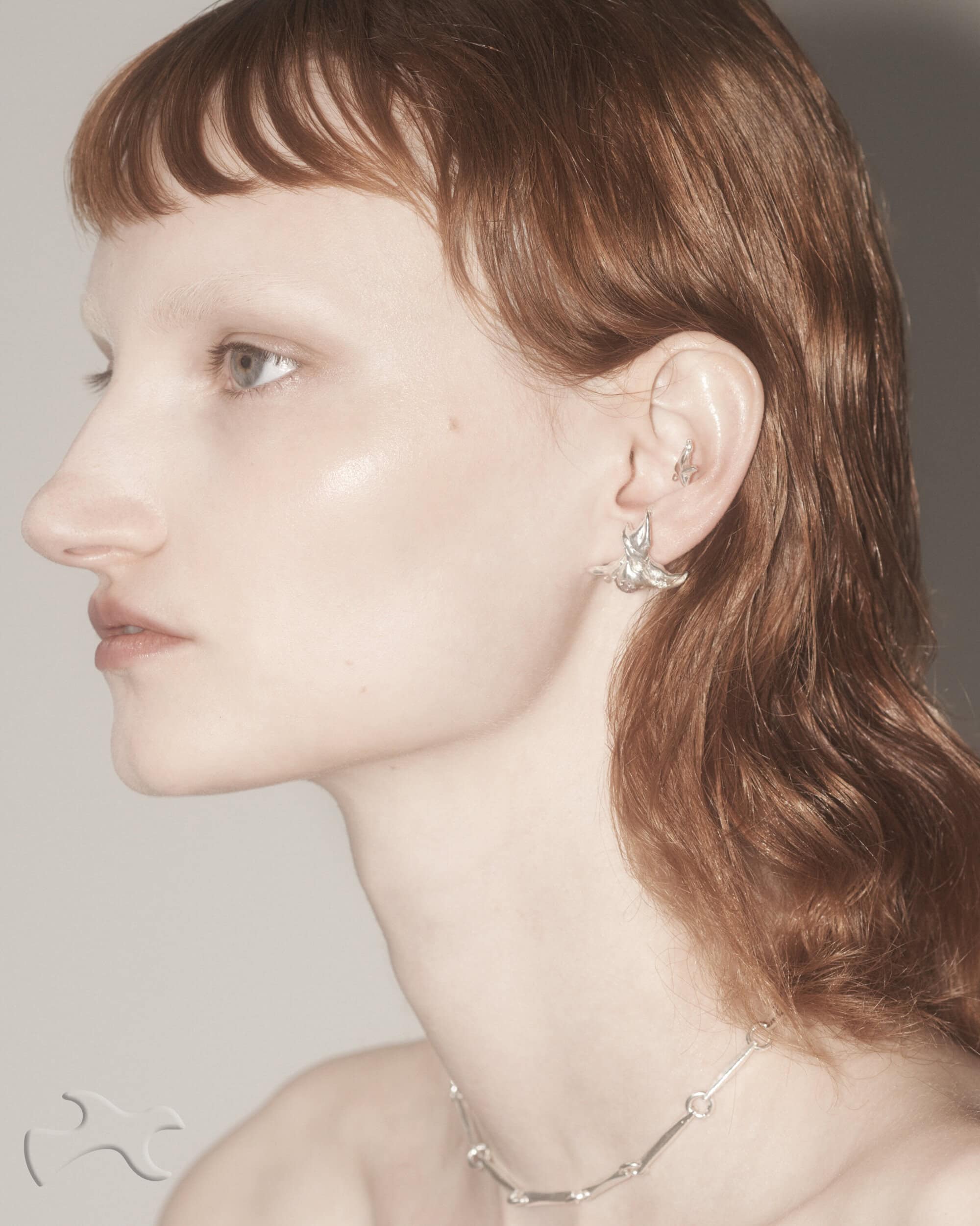 Close-up studio shot of a young adult female with auburn hair and pale skin wearing silver earrings and a necklace against a neutral background.