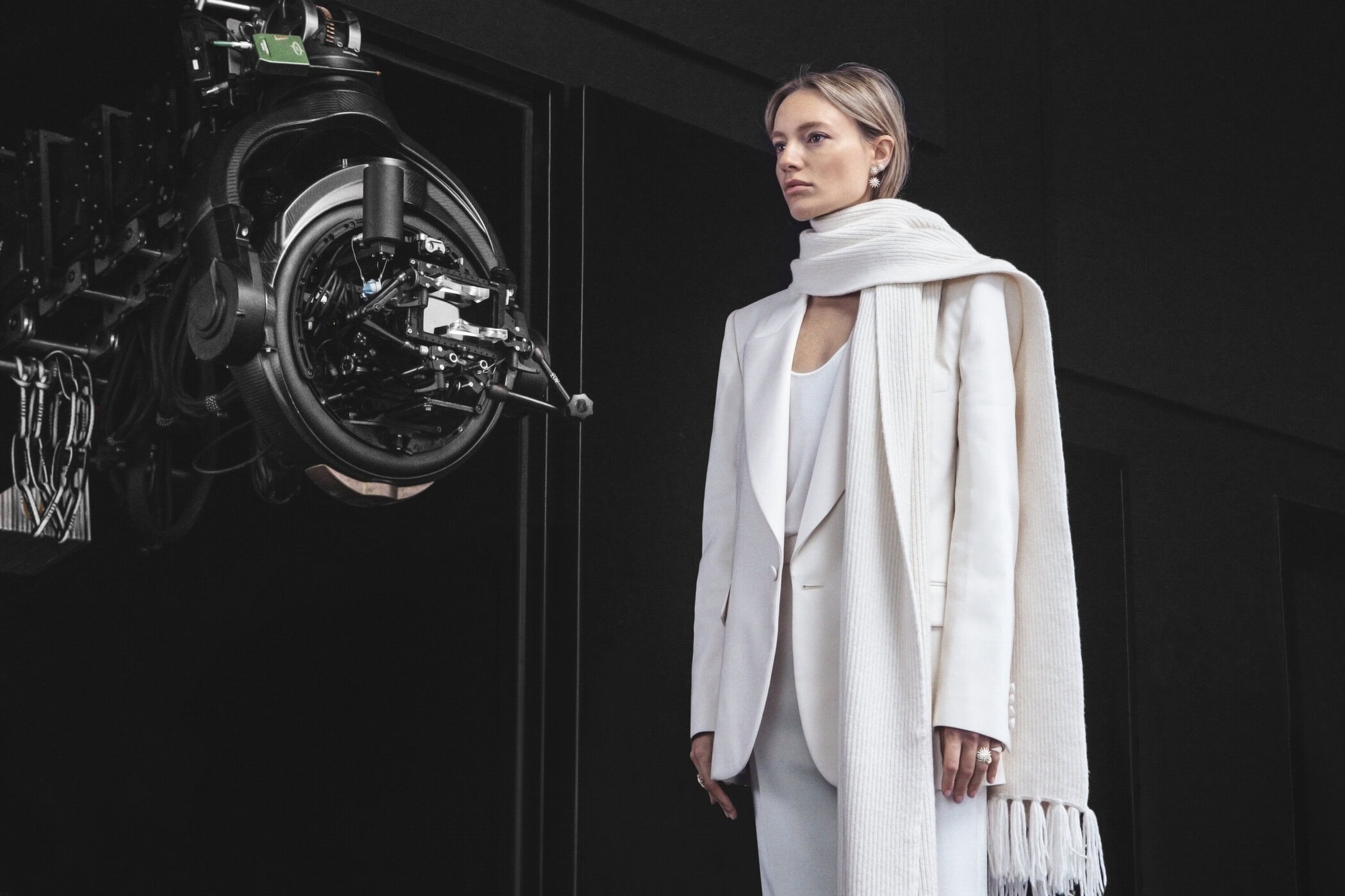 Fashion shot of an adult woman in a white suit and scarf standing in front of a large camera on a dark background indoors.