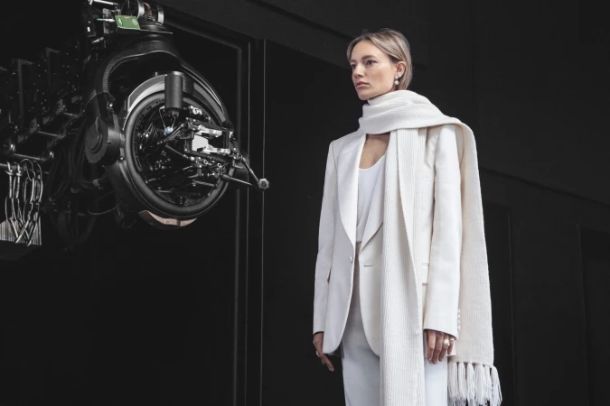 Fashion shot of an adult woman in a white suit and scarf standing in front of a large camera on a dark background indoors.