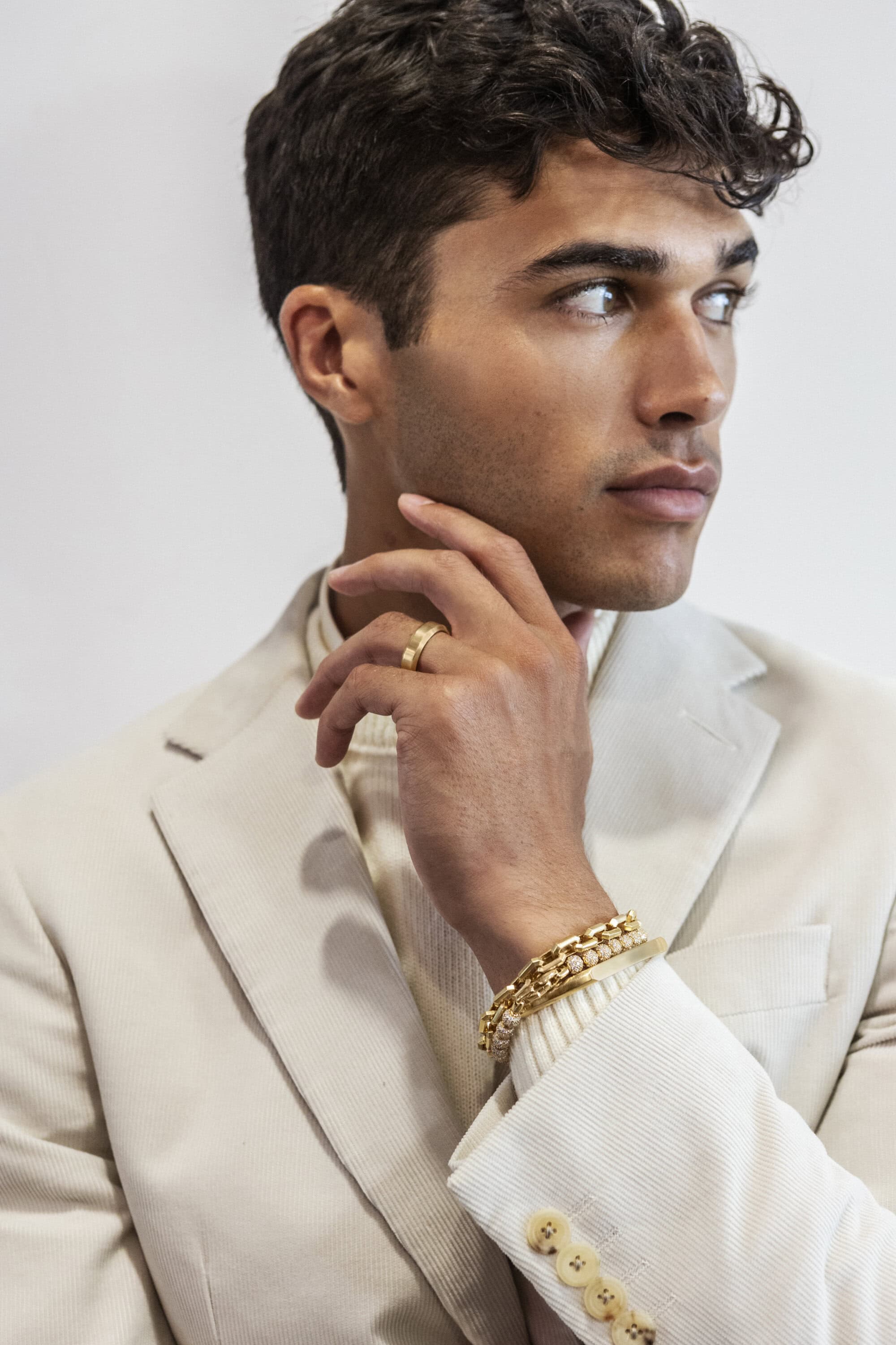 Portrait of a young adult male wearing a cream-colored suit, turtleneck sweater, and gold jewelry looking off to the side in a studio setting.