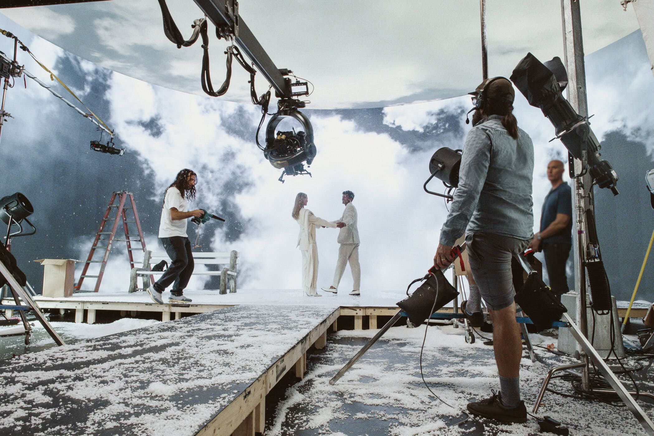 Film set with a couple in white holding hands, workers, camera crane, fake snow, and backdrop of clouds and storm, with stage lighting.