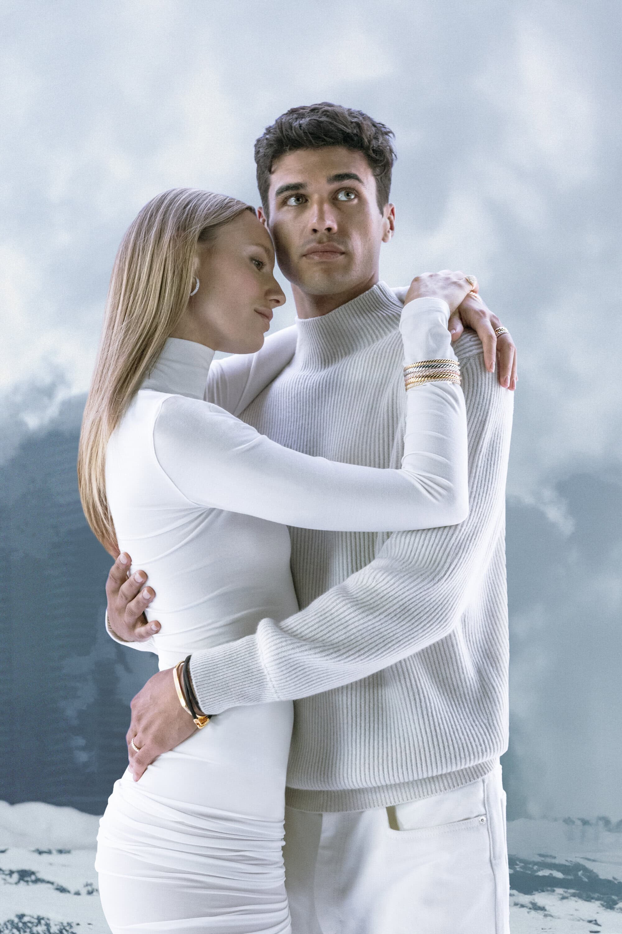 A fashionable young couple in white embraces outdoors against a diffused mountain backdrop, showcasing contemporary apparel and romantic mood.