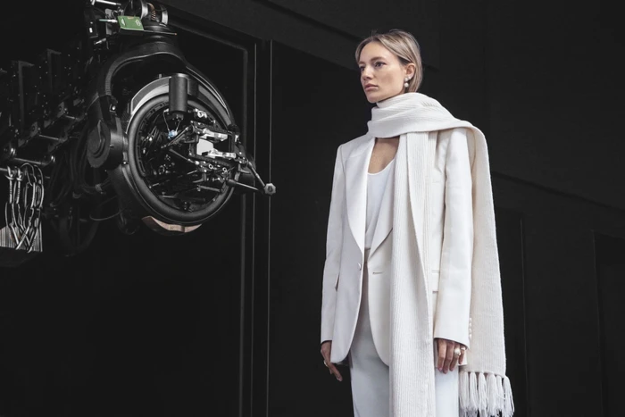Woman in white blazer and matching scarf standing beside professional camera equipment on a dark background