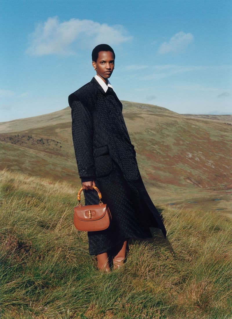 A slender, dark-skinned woman poses outdoors in a stylish dark jacket, skirt, and tan boots, holding a brown purse against a hilly landscape.