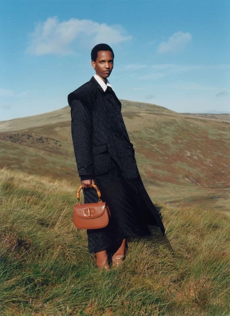 A slender, dark-skinned woman poses outdoors in a stylish dark jacket, skirt, and tan boots, holding a brown purse against a hilly landscape.