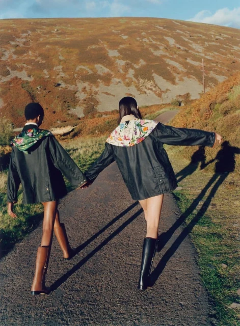 Two people walk along a path in the countryside, wearing matching dark green jackets and tall riding boots, with a large hill in the background.