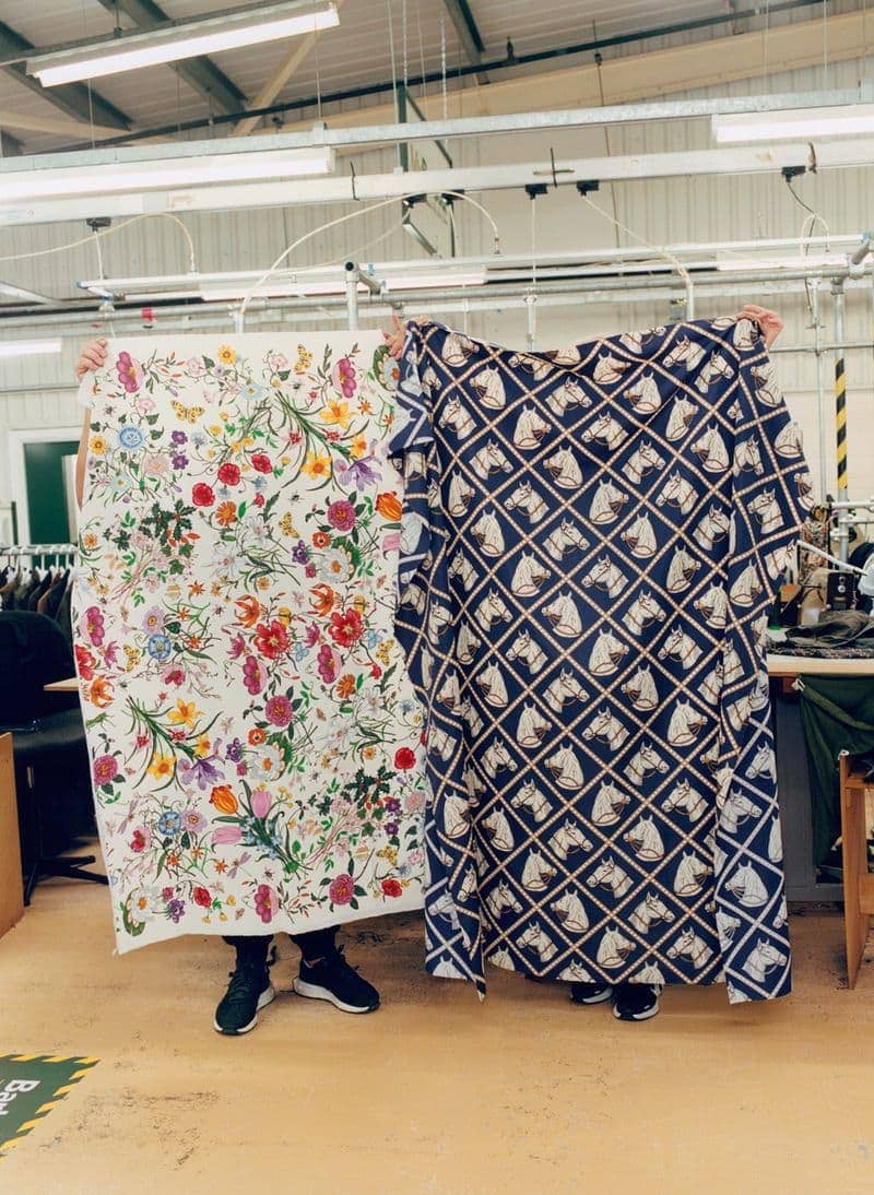 Two people holding up floral and horse-patterned fabrics indoors. Clothing factory with fluorescent lighting and industrial atmosphere, neutral background.