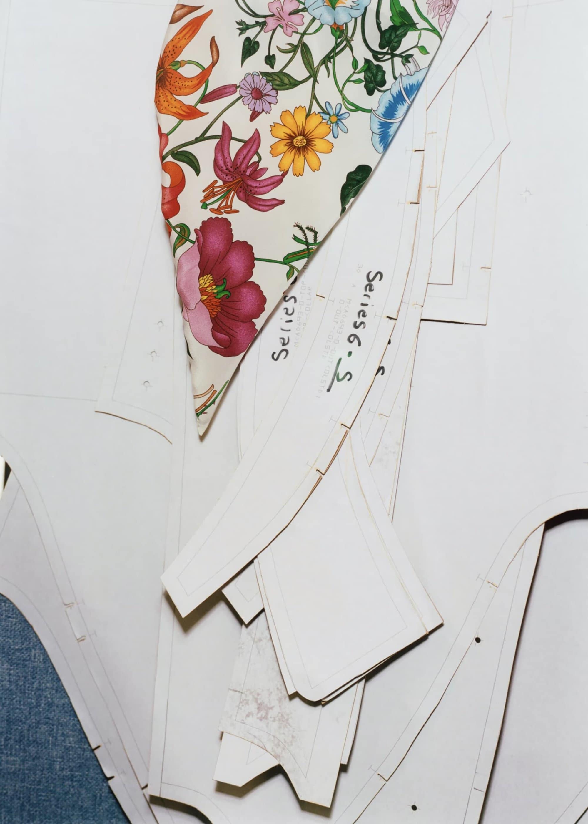 Floral fabric with pink, orange, and yellow flowers sits atop white paper sewing patterns on a white surface in a studio setting.
