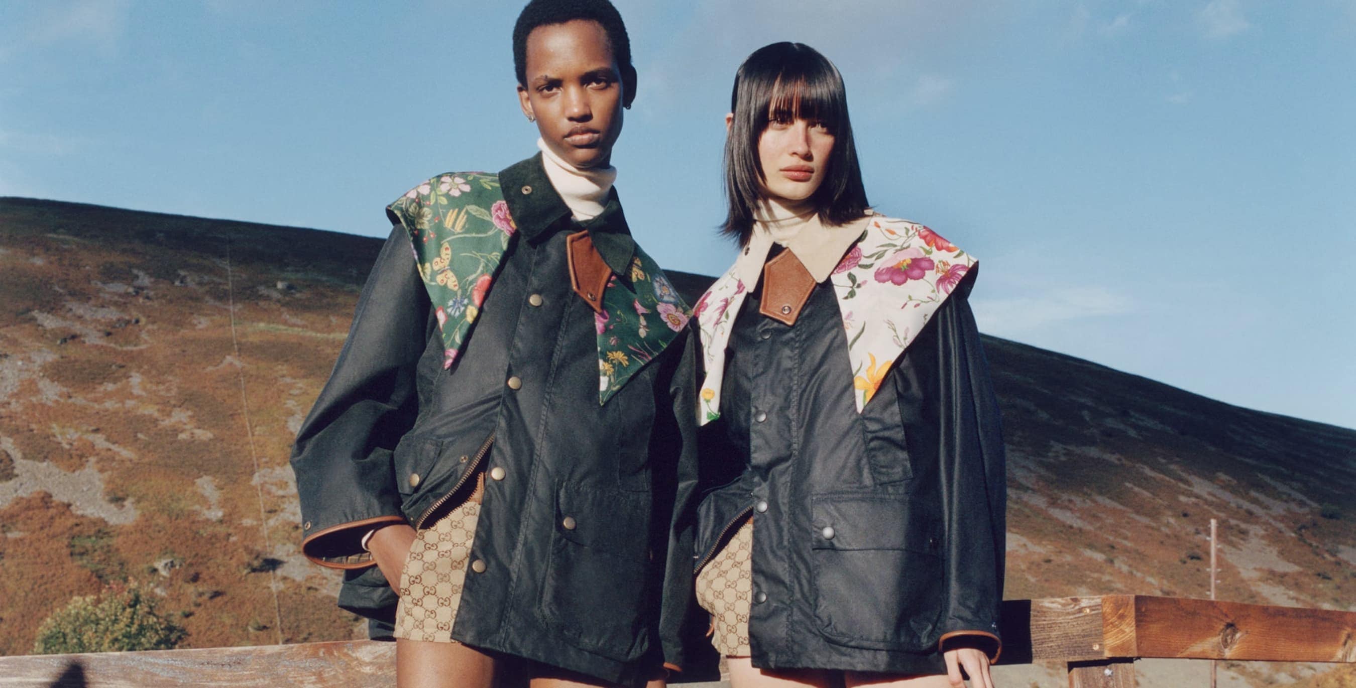 Two people wearing patterned jackets pose in front of a hill and blue sky. The jackets have a floral pattern collar.