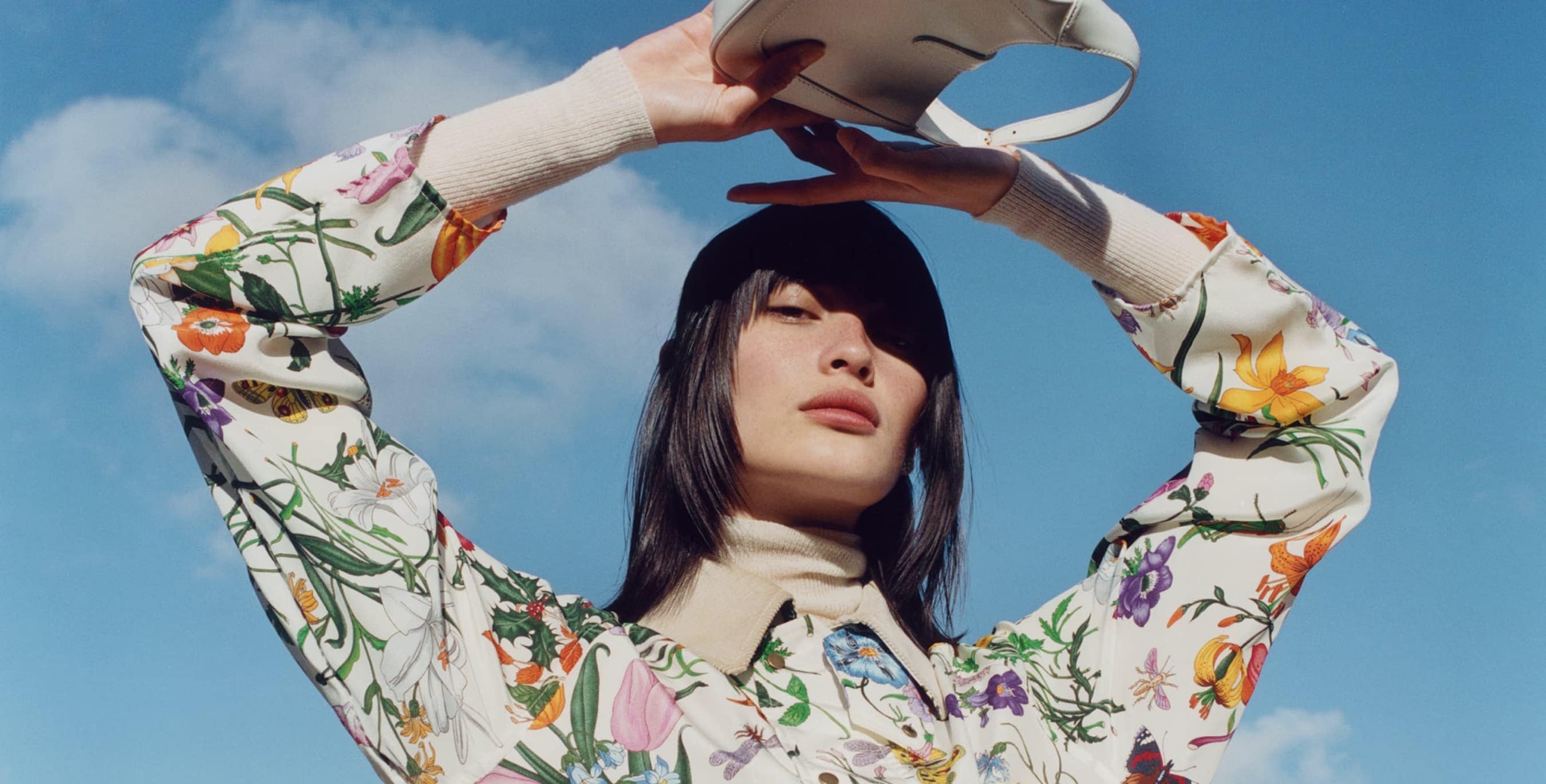 Woman in floral shirt holds white purse against blue sky. Fashion shot with natural light, soft colors, and a focus on spring style.