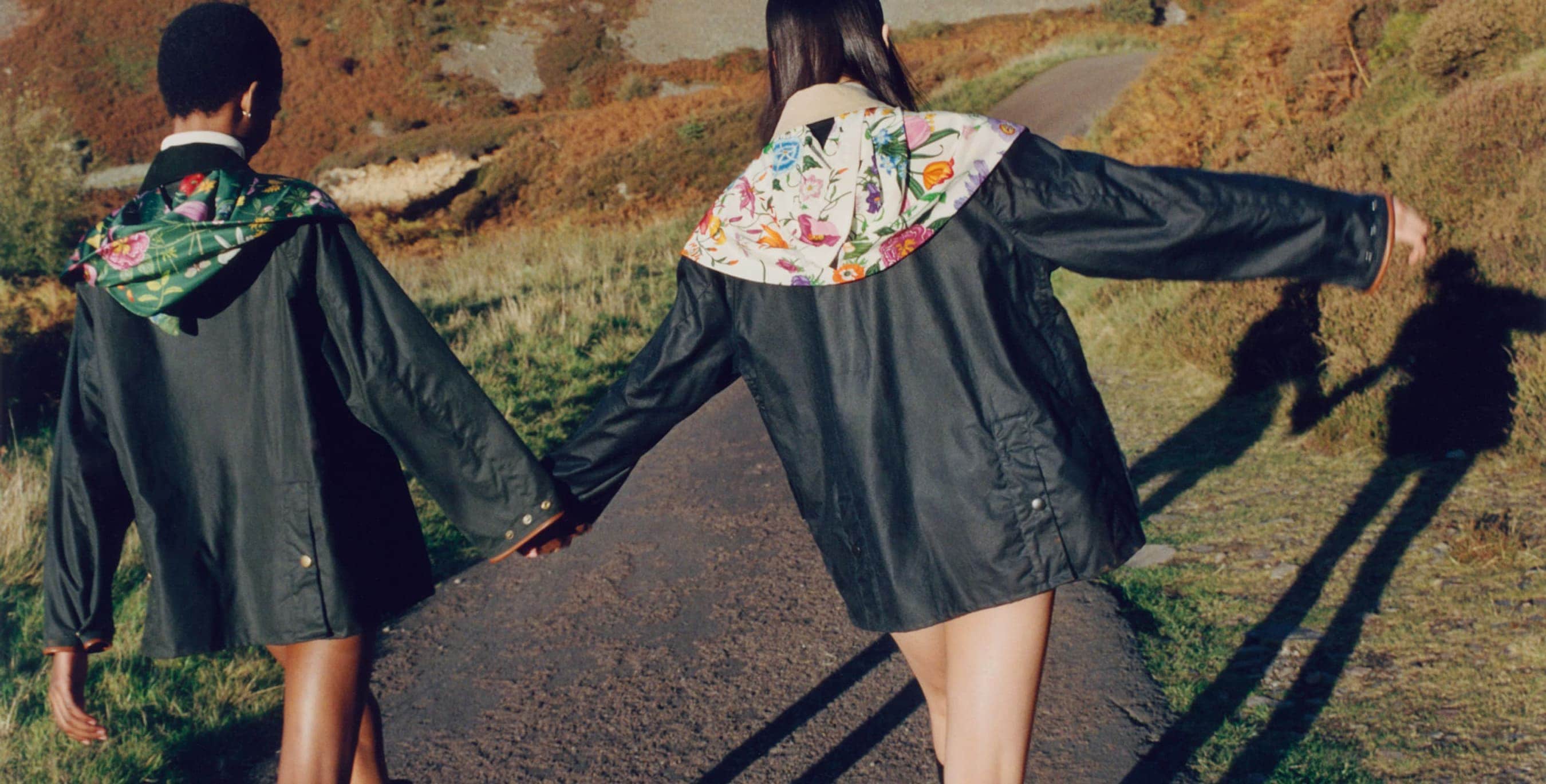 Two women walk on a path wearing matching black jackets with floral hoods in a rural landscape; their long shadows stretch before them.