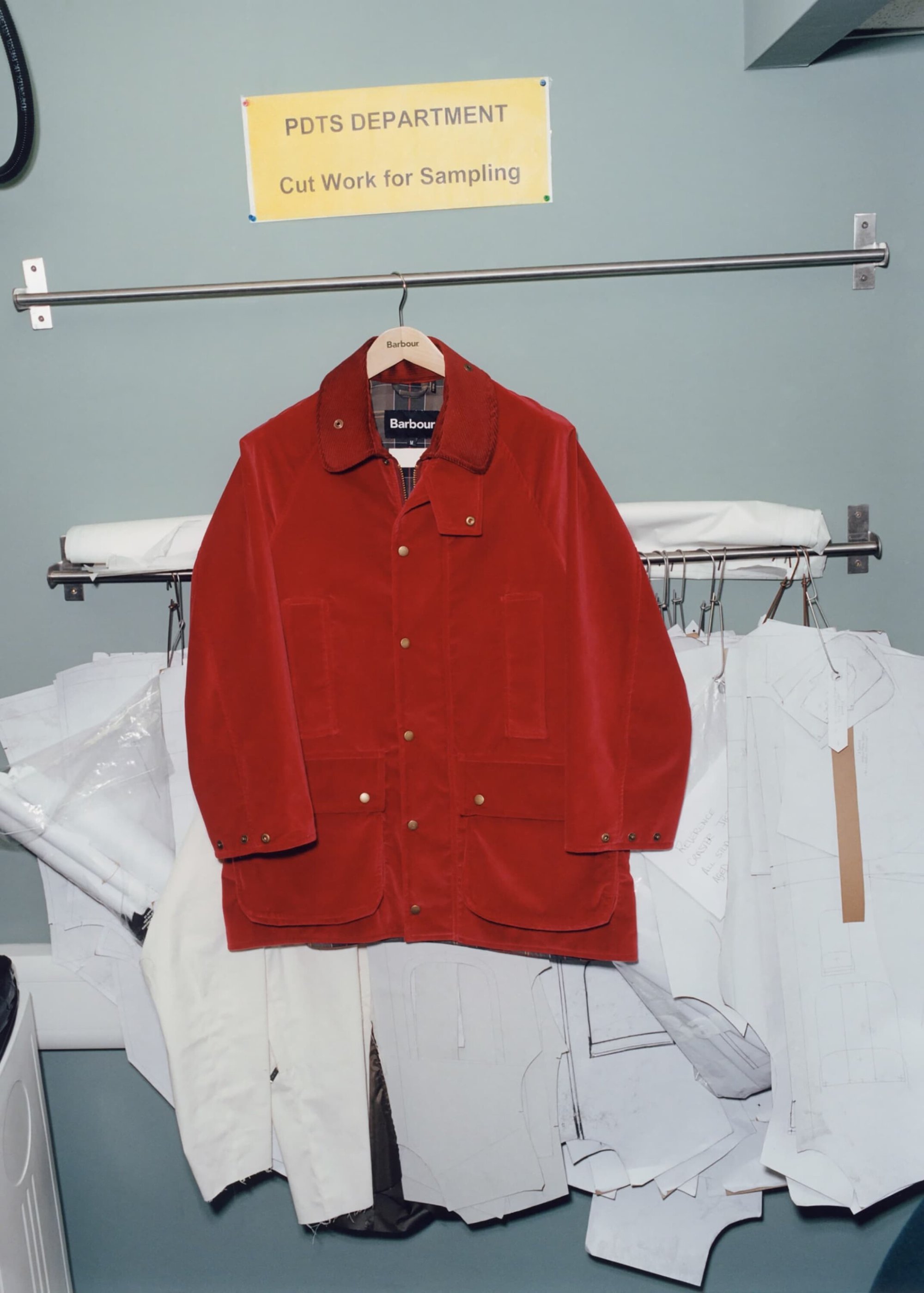 Red Barbour jacket hangs on a clothing rack surrounded by white fabric samples in a PDTS department room, displayed in a bright, natural light.