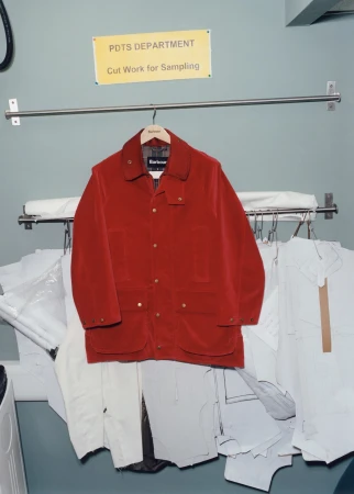 Red Barbour jacket hangs on a clothing rack surrounded by white fabric samples in a PDTS department room, displayed in a bright, natural light.
