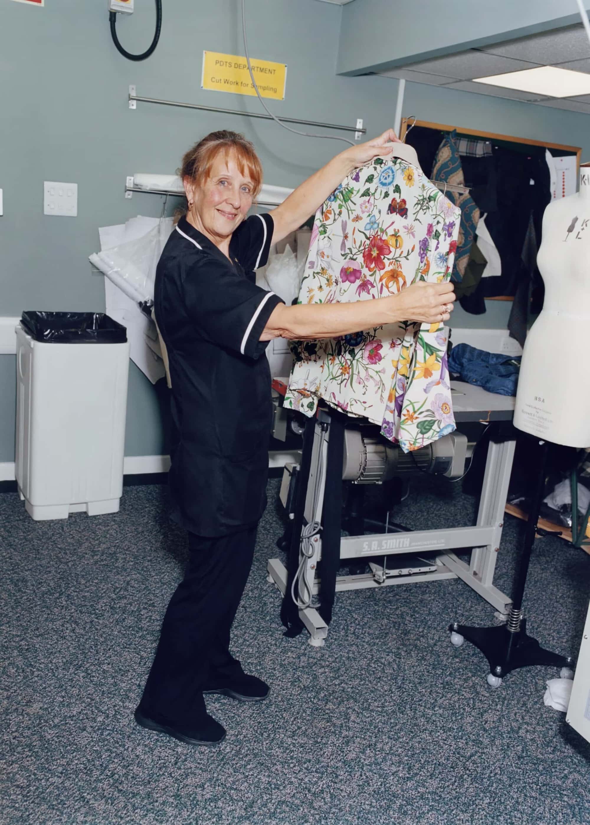 Mature adult female displays a floral jacket on a hanger in an indoor tailoring shop setting. Dress form and sewing machine visible.