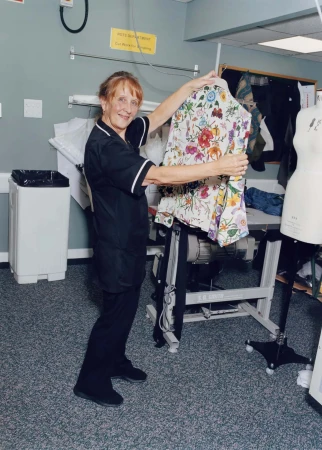Mature adult female displays a floral jacket on a hanger in an indoor tailoring shop setting. Dress form and sewing machine visible.