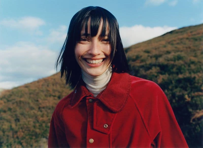 Smiling woman with dark hair in red jacket poses against a background of grassy hills and bright blue sky with scattered clouds.