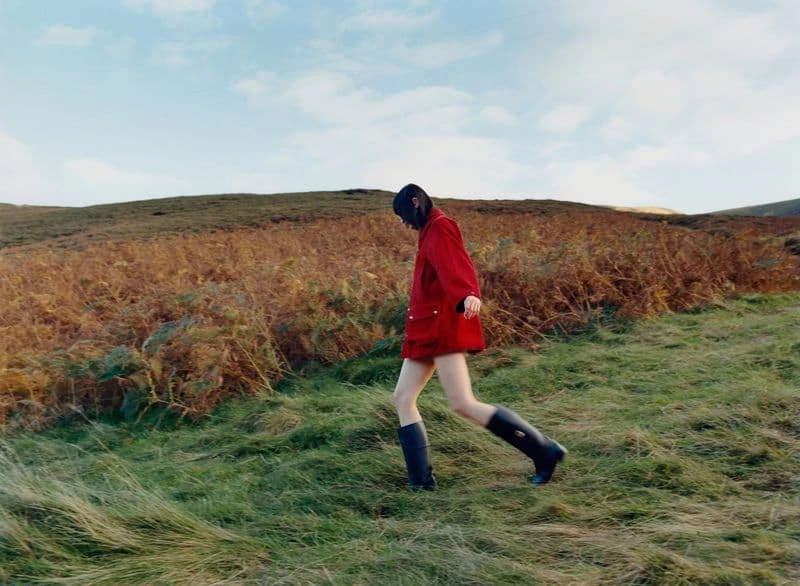 Woman in red coat and rubber boots walks through grassy field on a sunny day with a hill in the background. Nature photography.