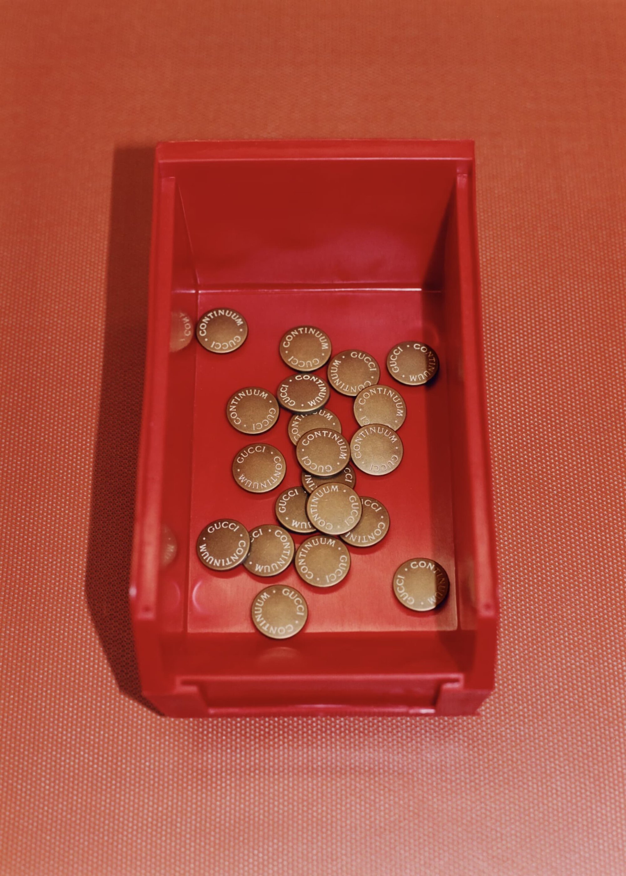 Red plastic container filled with gold-colored metal tokens featuring embossed 'Gucci' and 'Continuum' lettering; close-up studio shot.